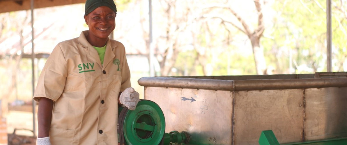 Ghanaian woman smiling and wearing brown SFC coat standing next to green motorized roaster underneath a shaded area