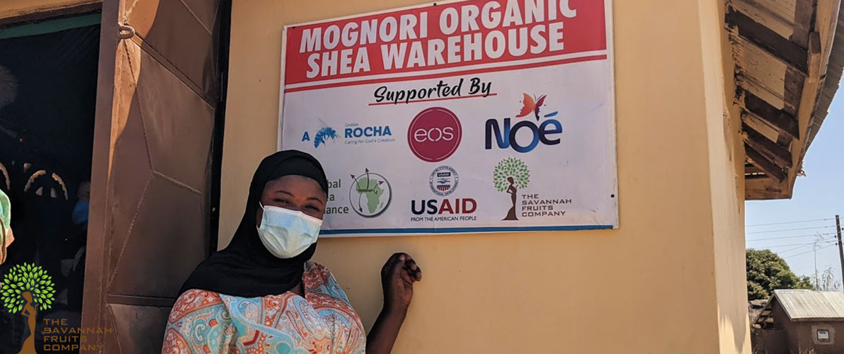 Ghanaian woman pointing to sign board hanging on outside of warehouse wall. Sign reads: Mognori Organic Shea Nuts Warehouse