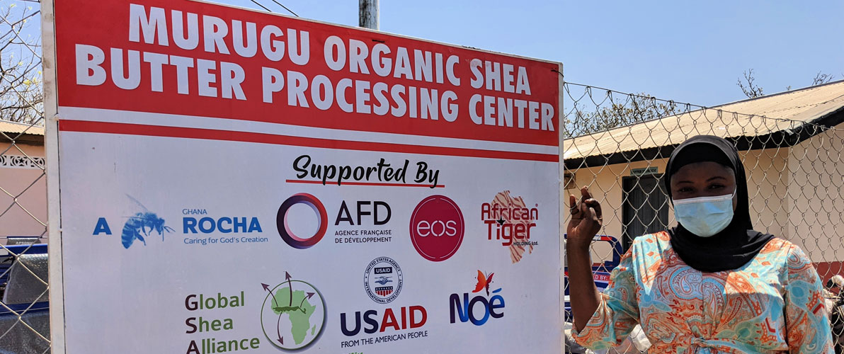 Large white cardboard sign board on metal chain link fence with signage “Murugu Organic Shea Butter Processing Center supported by” with logos of ten funders and partners below”. Ghanaian woman standing to the right of sign